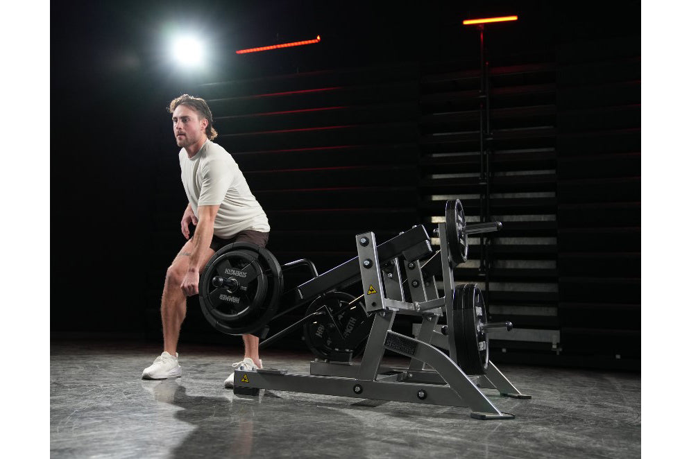 Man performing high pull exercise on black plate-loaded Hammer Strength machine in gym setting with neon lighting