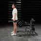 Man in cream t-shirt and brown shorts performing squat exercise on black Hammer Strength plate-loaded machine in gym setting