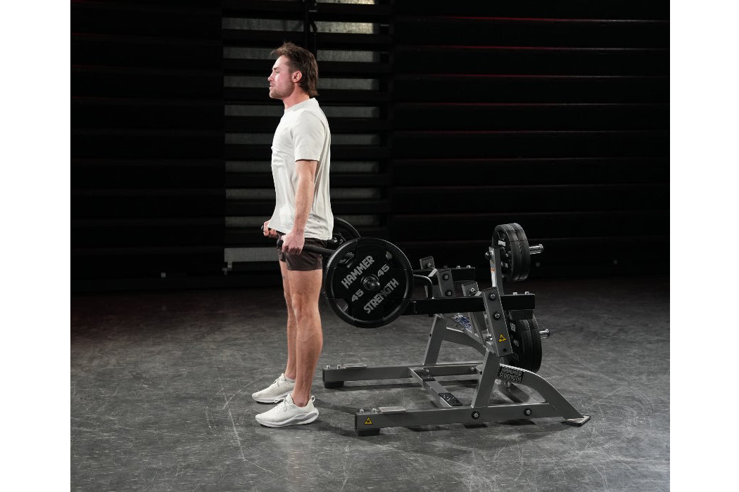 Man in cream t-shirt and brown shorts performing squat exercise on black Hammer Strength plate-loaded machine in gym setting