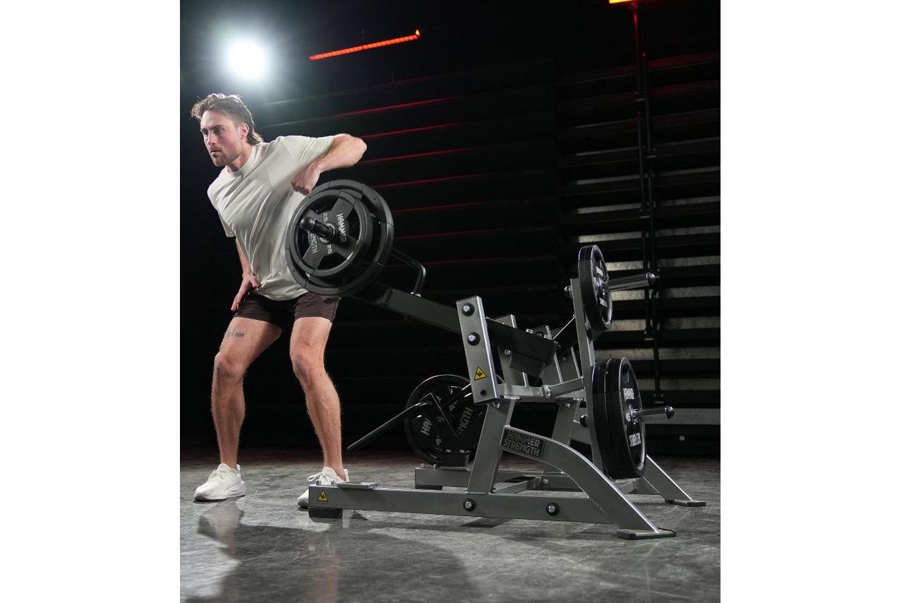 Man wearing light gray t-shirt and black shorts performing squat high pull exercise on Hammer Strength plate-loaded machine i