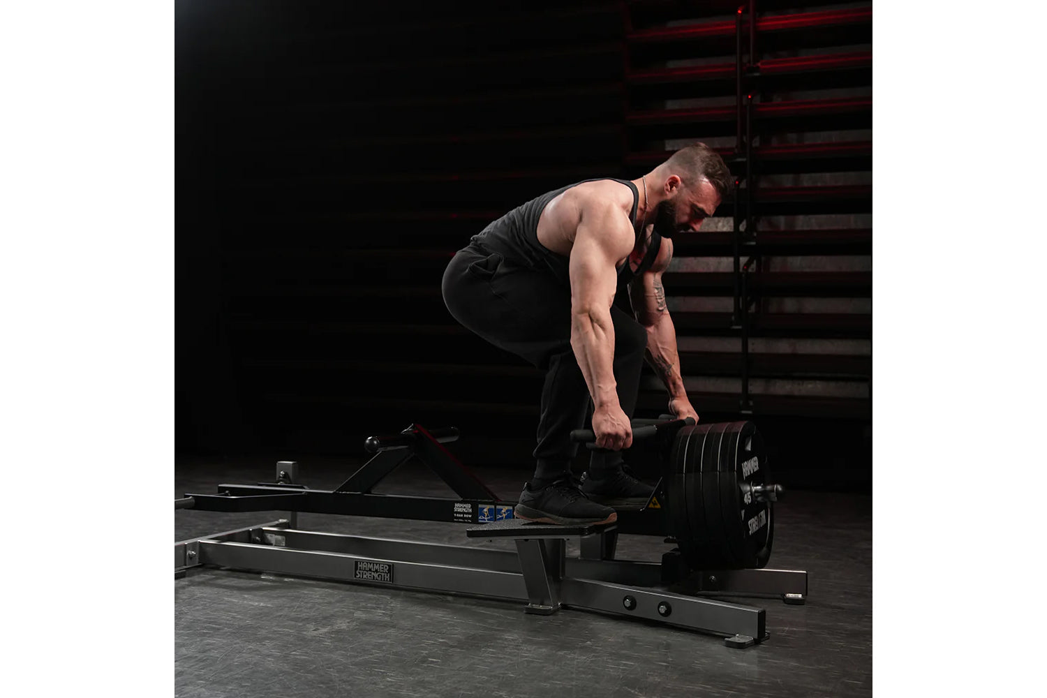 Man performing bent-over rowing exercise on black plate-loaded T-bar machine in gym setting with metal stairs background
