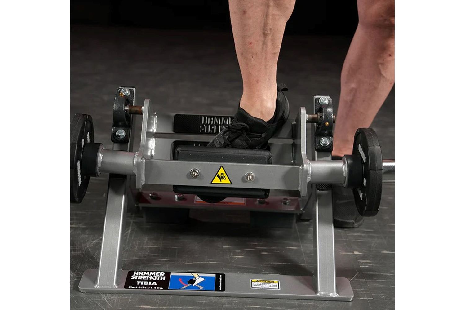 Person in black athletic shoe using Hammer Strength plate-loaded tibia dorsi flexion machine with gray frame and black footpl