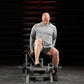 Man using plate-loaded tibia dorsi flexion bench machine in gym setting, wearing gray long-sleeve shirt and charcoal shorts