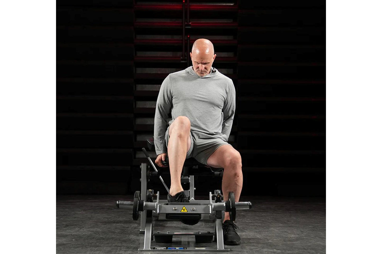 Man using plate-loaded tibia dorsi flexion bench machine in gym setting, wearing gray long-sleeve shirt and charcoal shorts