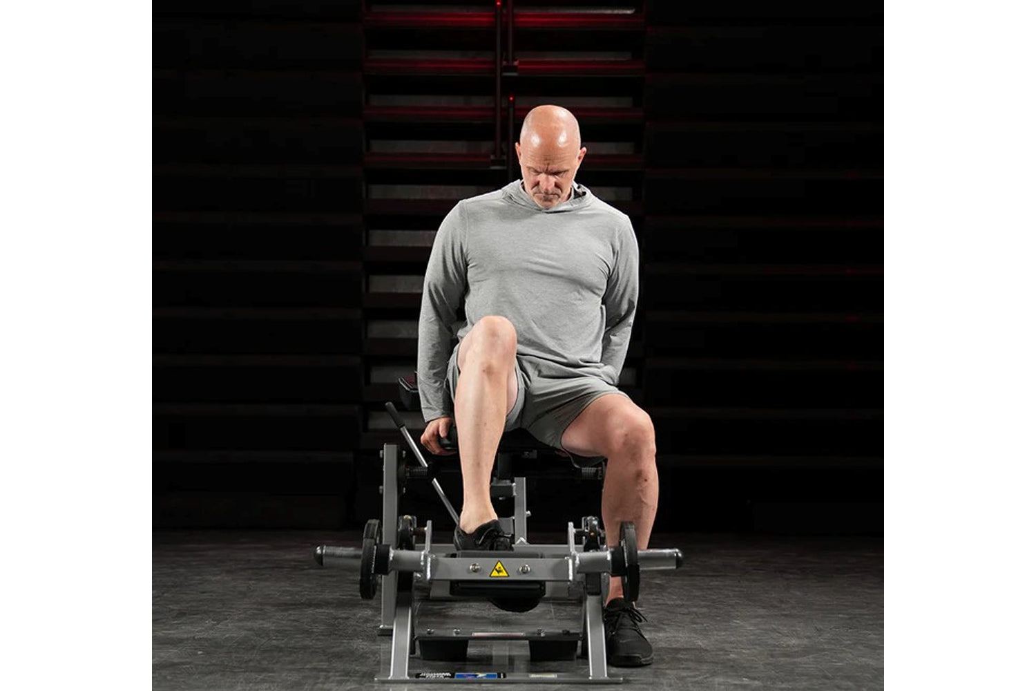 Man using plate-loaded tibia dorsi flexion bench machine in gym setting, wearing gray long-sleeve shirt and charcoal shorts