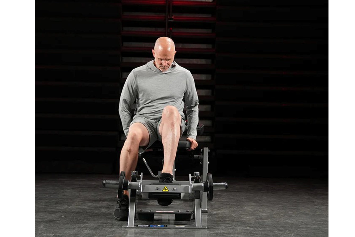 Male model on leg flexion machine wearing gray long-sleeve shirt and shorts, demonstrating exercise position in gym setting