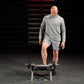 Man wearing gray long-sleeve shirt and shorts standing on plate-loaded tibia dorsi flexion exercise machine in gym setting