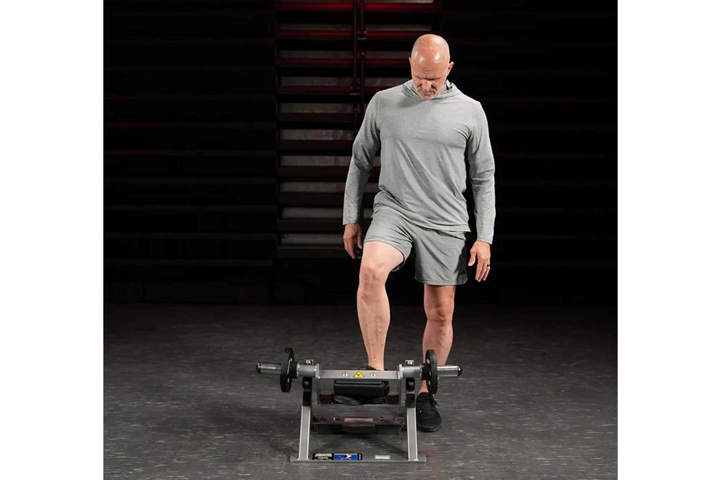 Man wearing gray long-sleeve shirt and shorts standing on plate-loaded tibia dorsi flexion exercise machine in gym setting