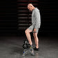 Man wearing gray Adidas hoodie and shorts using plate-loaded tibia dorsi flexion bench machine in gym setting