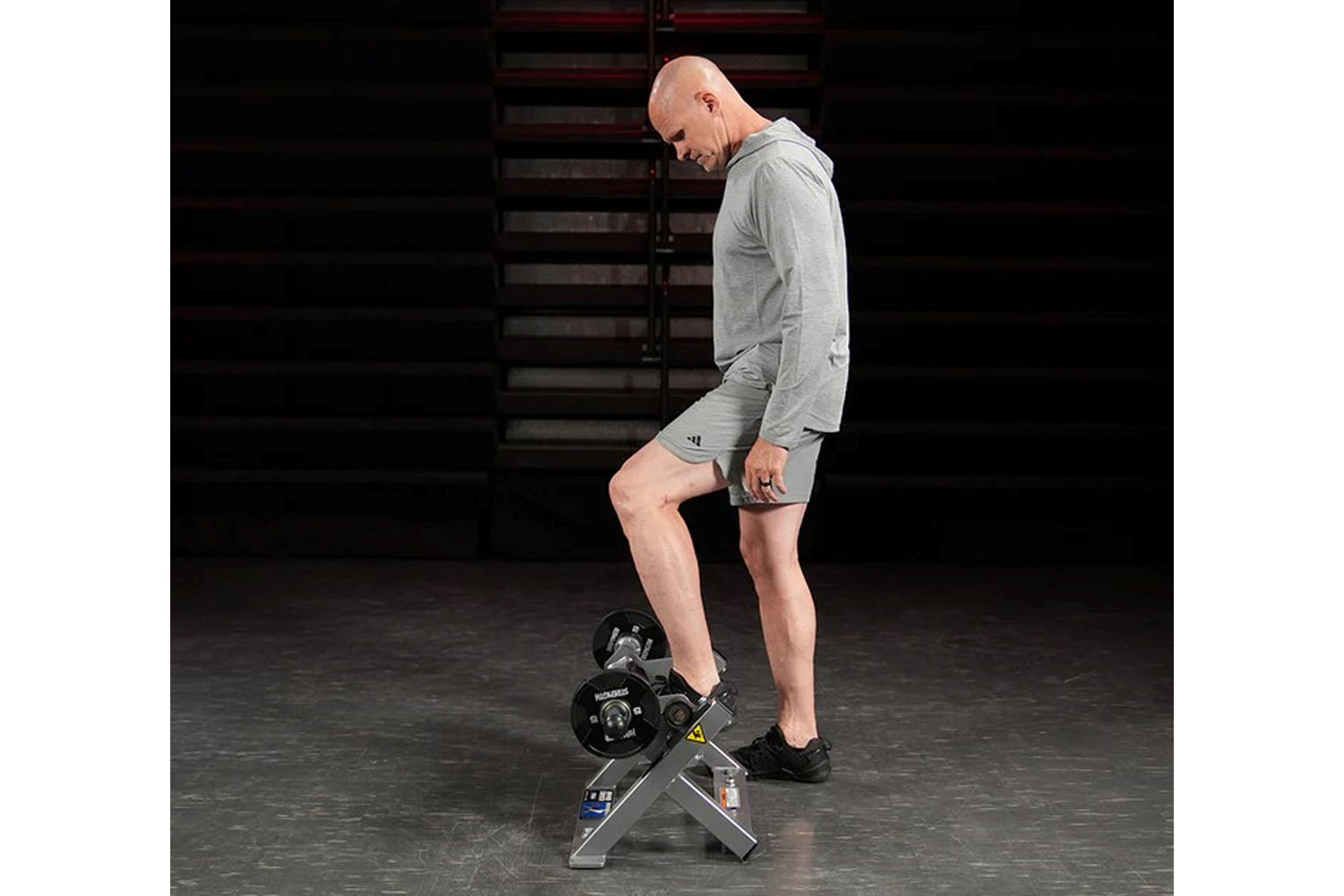 Man wearing gray Adidas hoodie and shorts using plate-loaded tibia dorsi flexion bench machine in gym setting