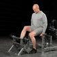 Man in gray long-sleeve shirt and shorts using plate-loaded tibia dorsi flexion machine with black weights in gym setting