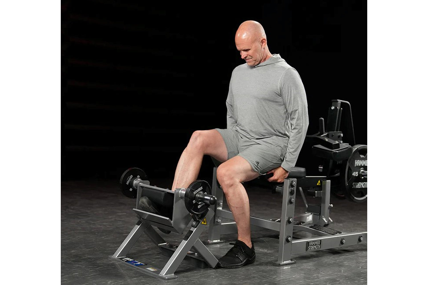 Man in gray long-sleeve shirt and shorts using plate-loaded tibia dorsi flexion machine with black weights in gym setting