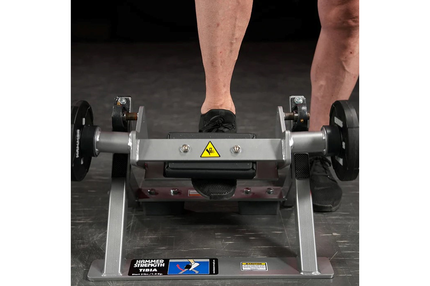 Man's feet on black footplate of Hammer Strength plate-loaded tibia dorsi flexion machine with silver frame and black weight 