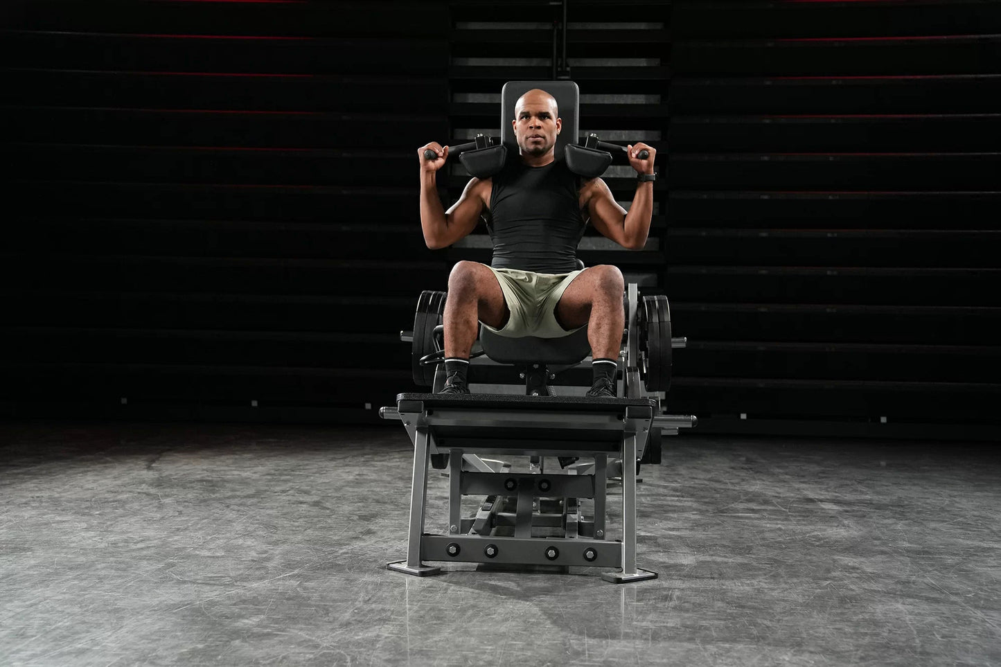 Man performing leg exercise on black plate-loaded V-squat machine with dumbbells, gym setting