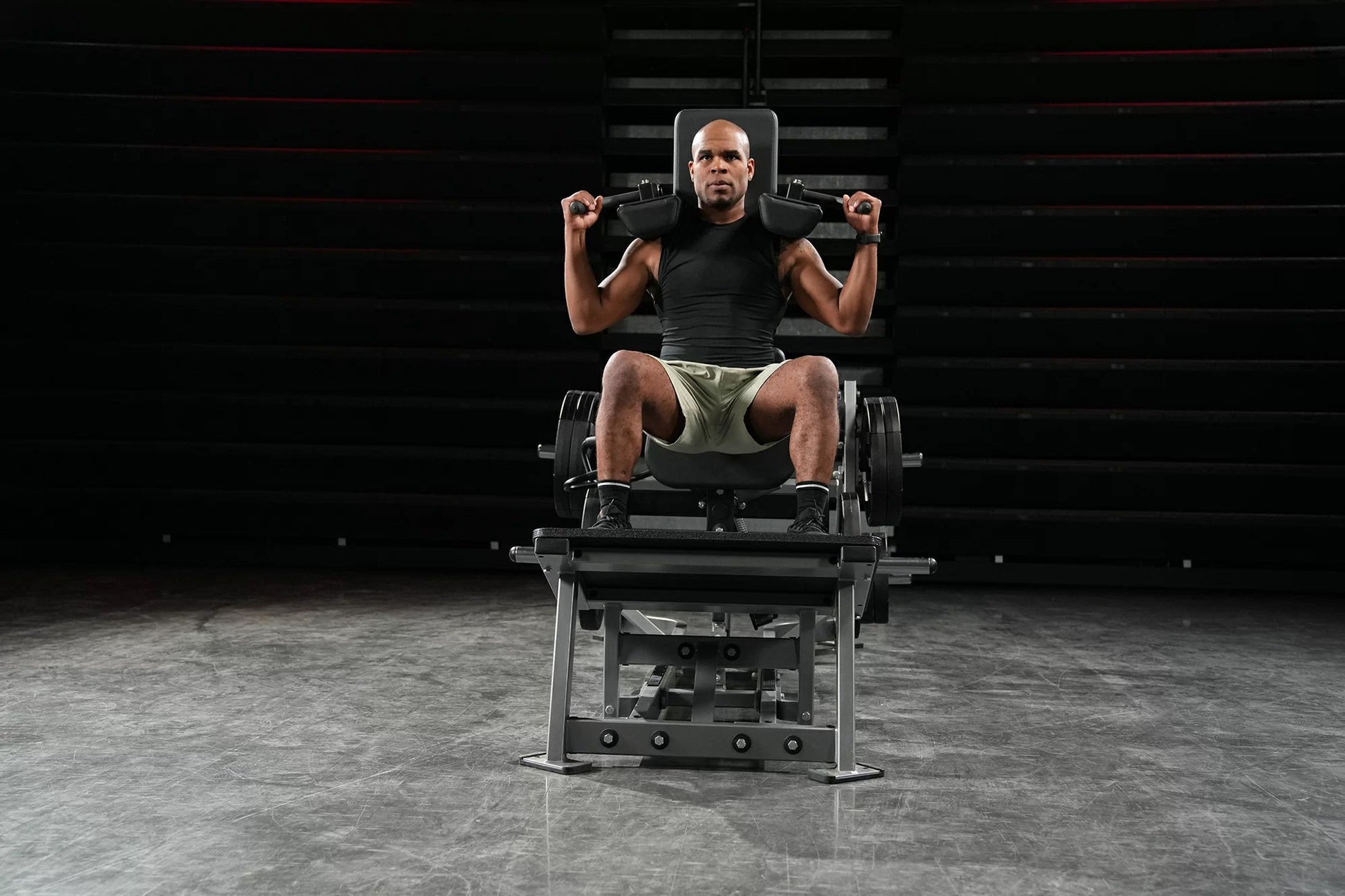 Man performing leg exercise on black plate-loaded V-squat machine with dumbbells, gym setting