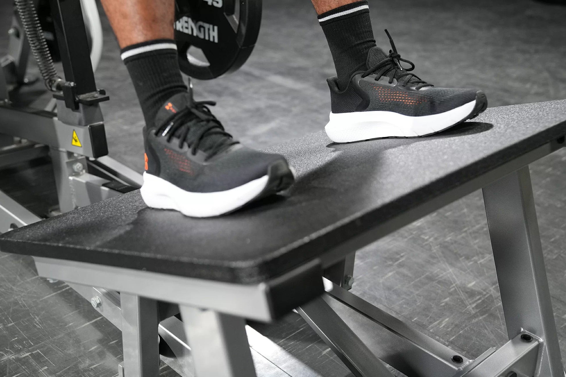 Black and white running shoes with orange accents worn by model on exercise equipment during workout
