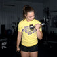 Woman wearing yellow graphic tee and black shorts lifting chrome dumbbell in gym setting