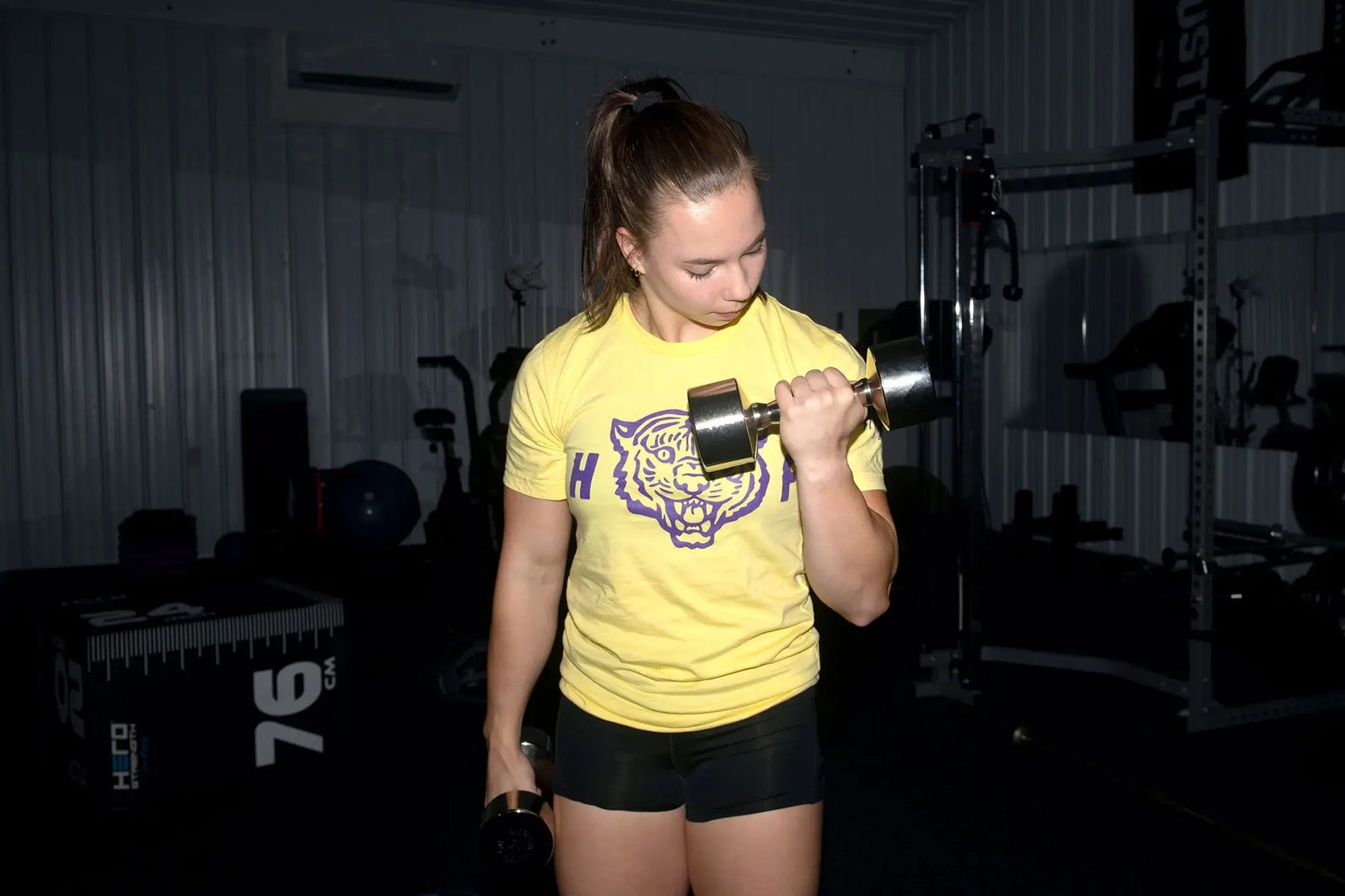Woman wearing yellow graphic tee and black shorts lifting chrome dumbbell in gym setting