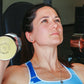 Woman performing dumbbell shoulder press with chrome dumbbells marked "Hampton 15" in blue and white sports bra, gym setting