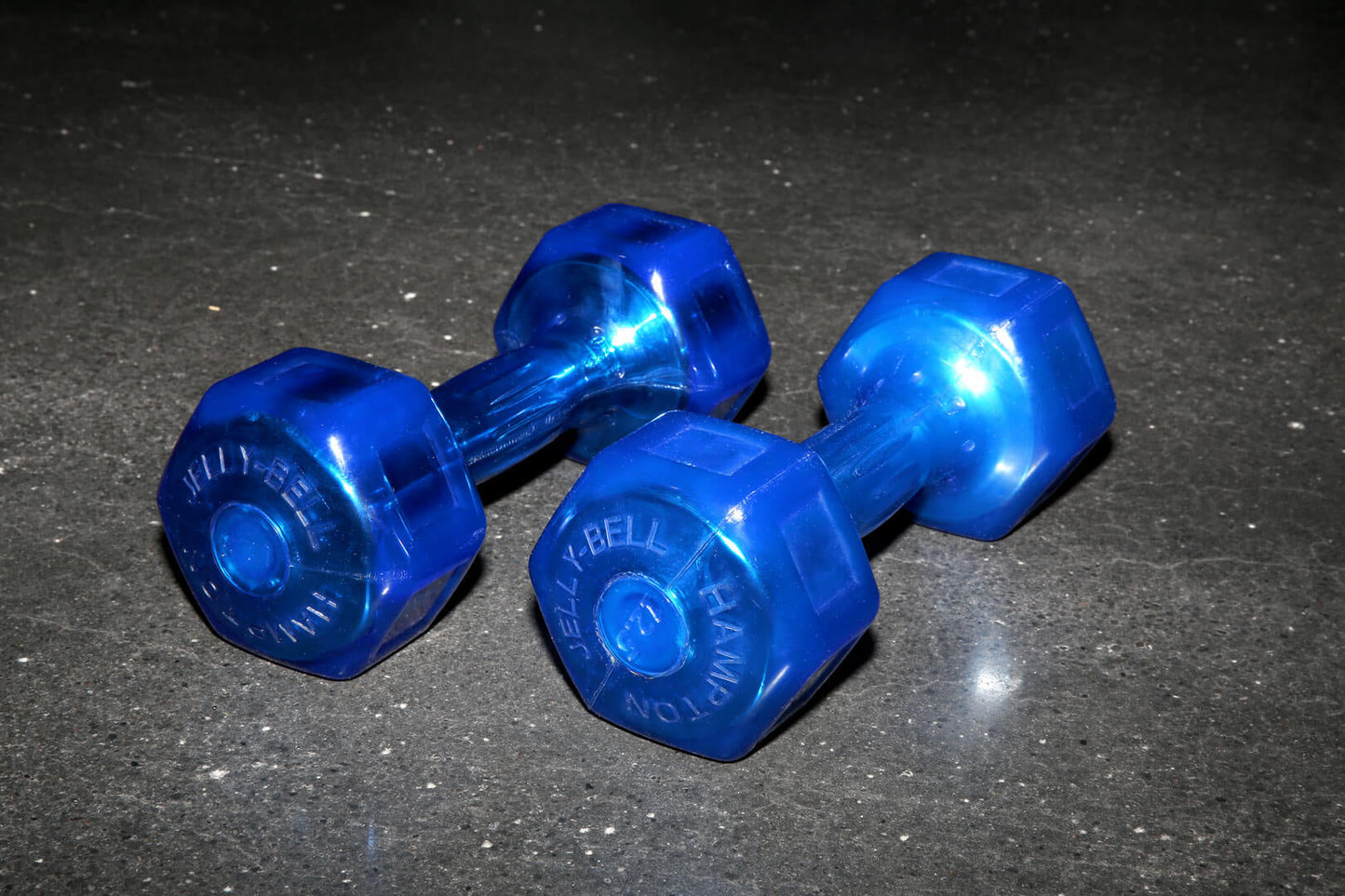 Pair of blue translucent hexagonal dumbbells with Jelly-Bell branding, displayed on dark textured surface