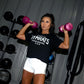 Woman wearing black "Hampton Gyrats" graphic tee and white shorts holding pink dumbbells in gym setting with equipment