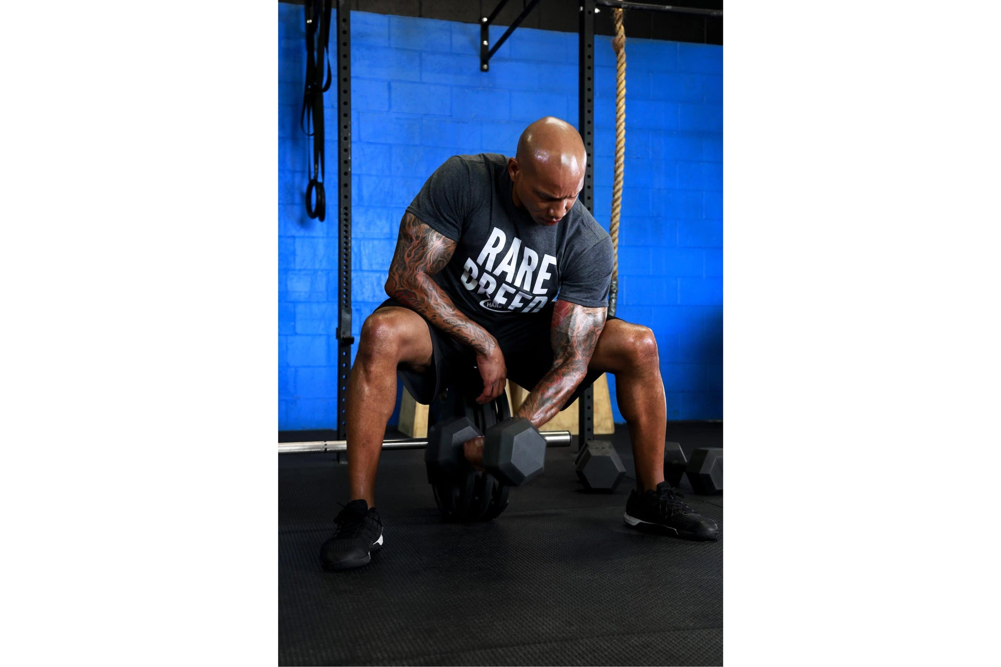 Man wearing black "Bare Speed" graphic t-shirt performing dumbbell exercise in gym with blue wall background