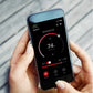 Smartphone displaying sauna control app with 74°C temperature dial, red steamer and fan controls, held in hand against white 