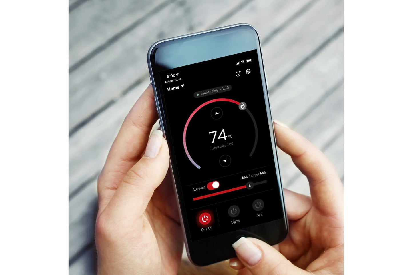 Smartphone displaying sauna control app with 74°C temperature dial, red steamer and fan controls, held in hand against white 