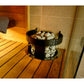 Black electric sauna heater with bowl of stones on top, positioned on wooden sauna bench with towel and shower area visible