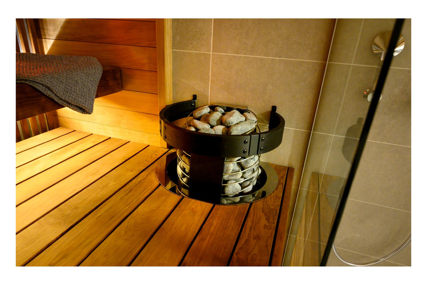 Black electric sauna heater with bowl of stones on top, positioned on wooden sauna bench with towel and shower area visible