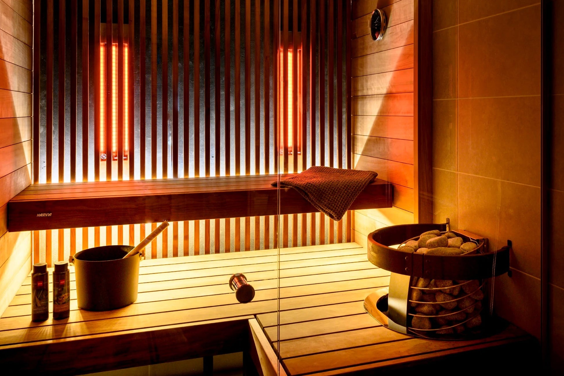 Modern sauna interior with Harvia Spirit heater, wooden benches, neon orange vertical lighting, bucket, ladle, and towel