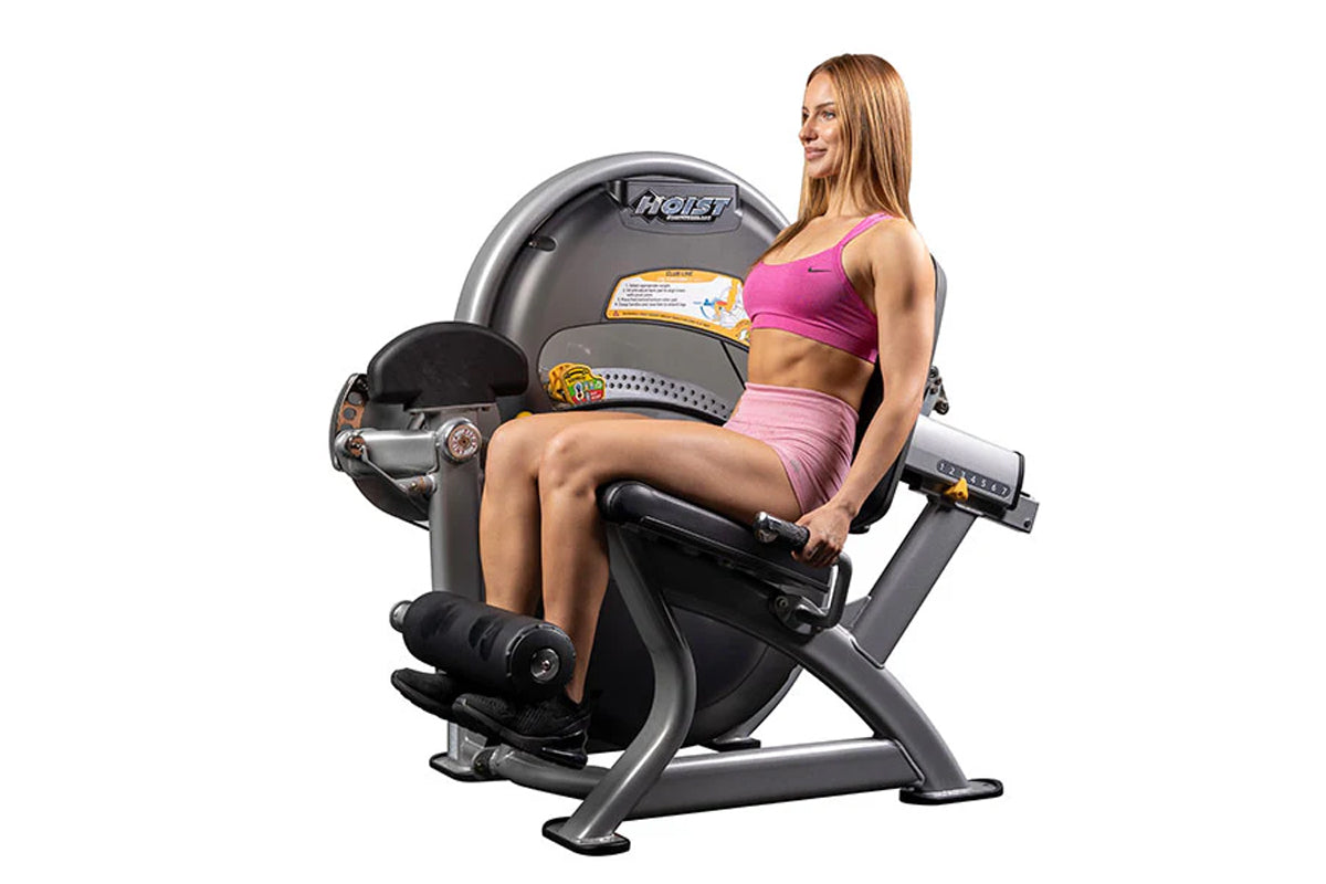 Woman in pink sports bra and shorts seated on Hoist leg extension machine with gray and black frame, shown on white backgroun