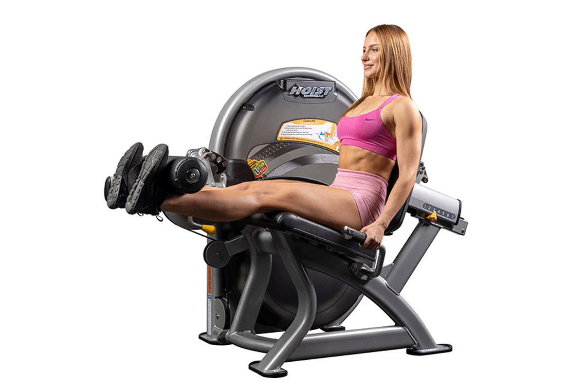 Woman in pink sports bra and shorts using black and gray Hoist leg extension machine with circular pad and foot platform, sho