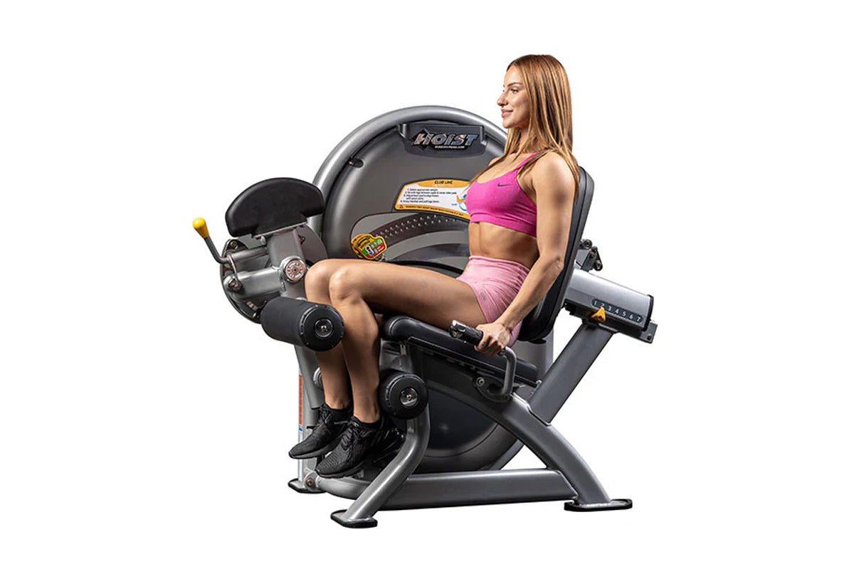Woman in pink sports bra and shorts seated on black and gray Hoist leg curl machine with padded seat and leg extension lever