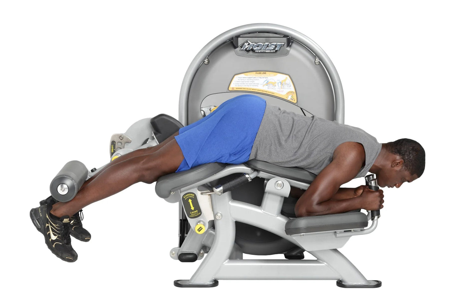 Man using Hoist Standing Prone Leg Curl machine, wearing blue shorts and gray shirt, on white background