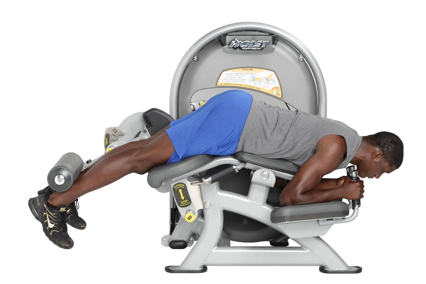 Man using Hoist Standing Prone Leg Curl machine, wearing blue shorts and gray shirt, on white background