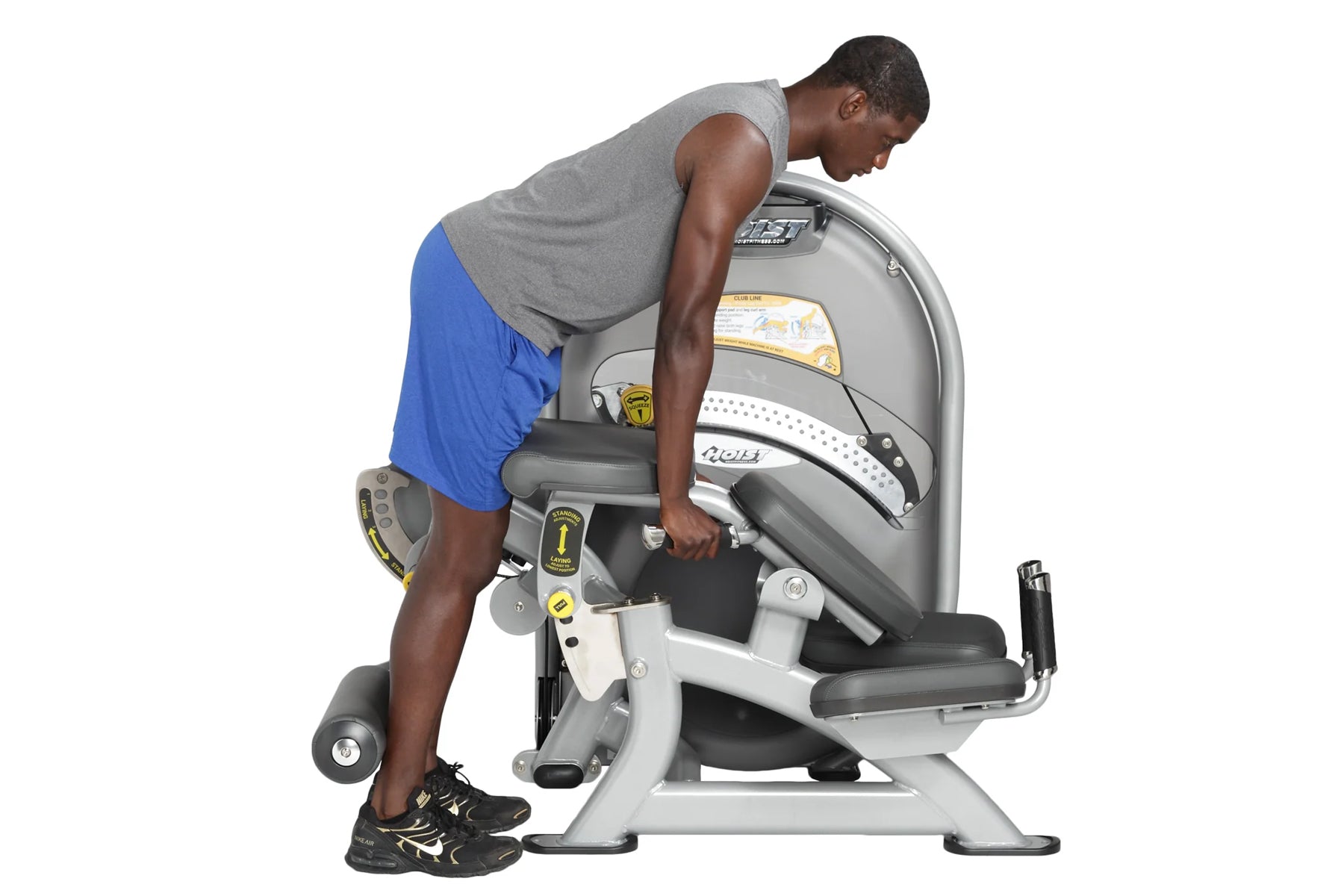 Man using gray and white Hoist Standing Prone Leg Curl machine with blue shorts and gray tank top, demonstrating leg exercise