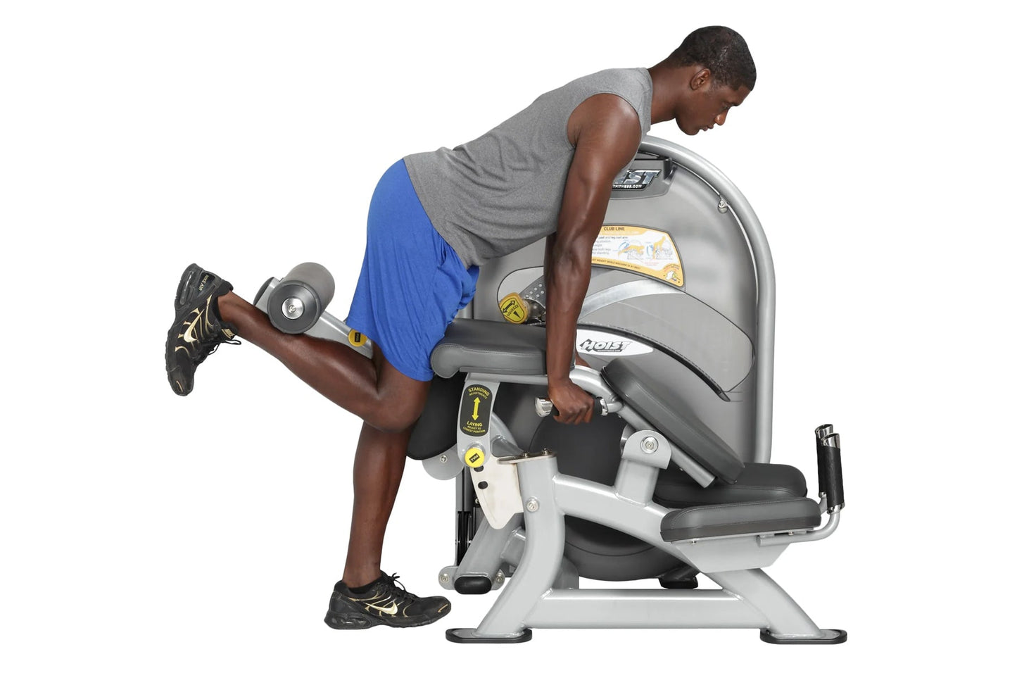 Man in gray tank top and blue shorts using white and gray leg curl machine with dumbbells on white background
