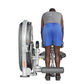 Man using silver and gray leg curl machine, wearing gray tank top and blue shorts, viewed from behind on white background