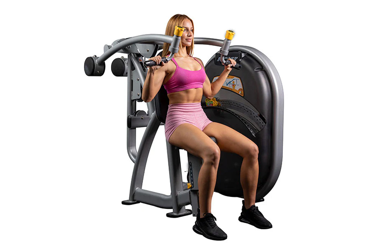 Woman using gray and black shoulder press machine with yellow handles, wearing magenta sports bra and pink shorts