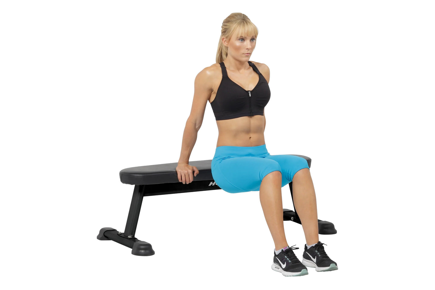 Black padded flat utility bench with sturdy frame, worn by model in black sports bra and blue shorts during exercise demonstr