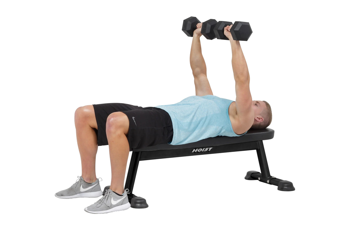 Man performing dumbbell chest press on black Hoist flat utility bench, wearing light blue shirt and black shorts