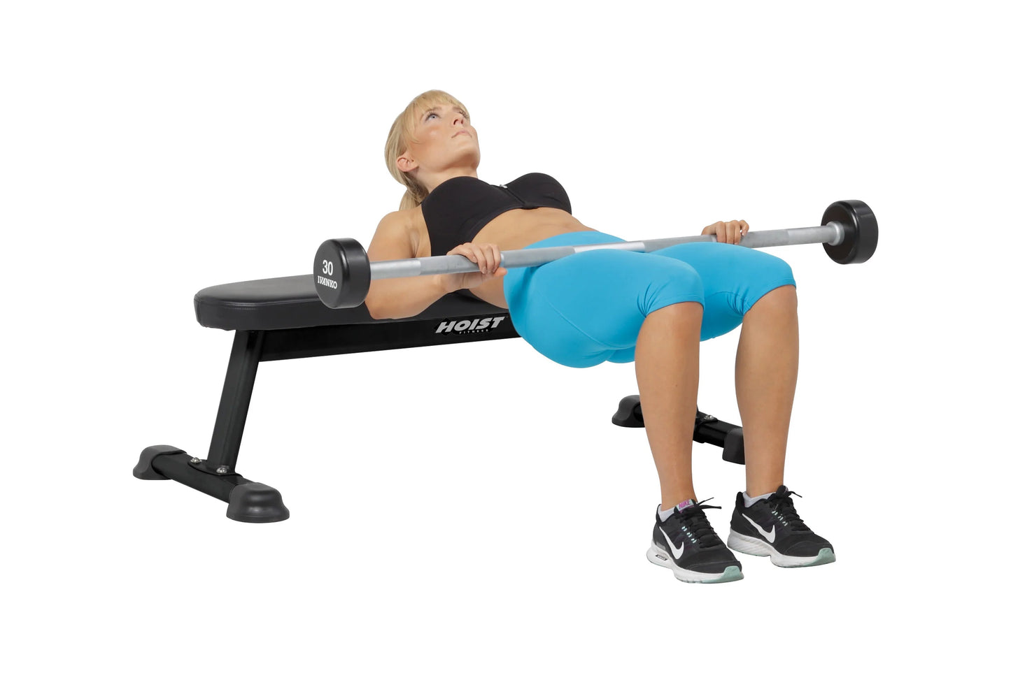 Woman performing barbell bench press on black Hoist flat utility bench with 30-pound dumbbells, wearing blue shorts and black