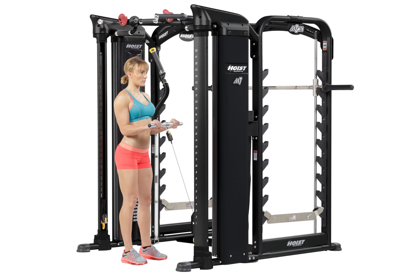 Woman in blue sports bra and coral shorts using Hoist MI7 black Smith machine with cable attachments and weight stack on whit