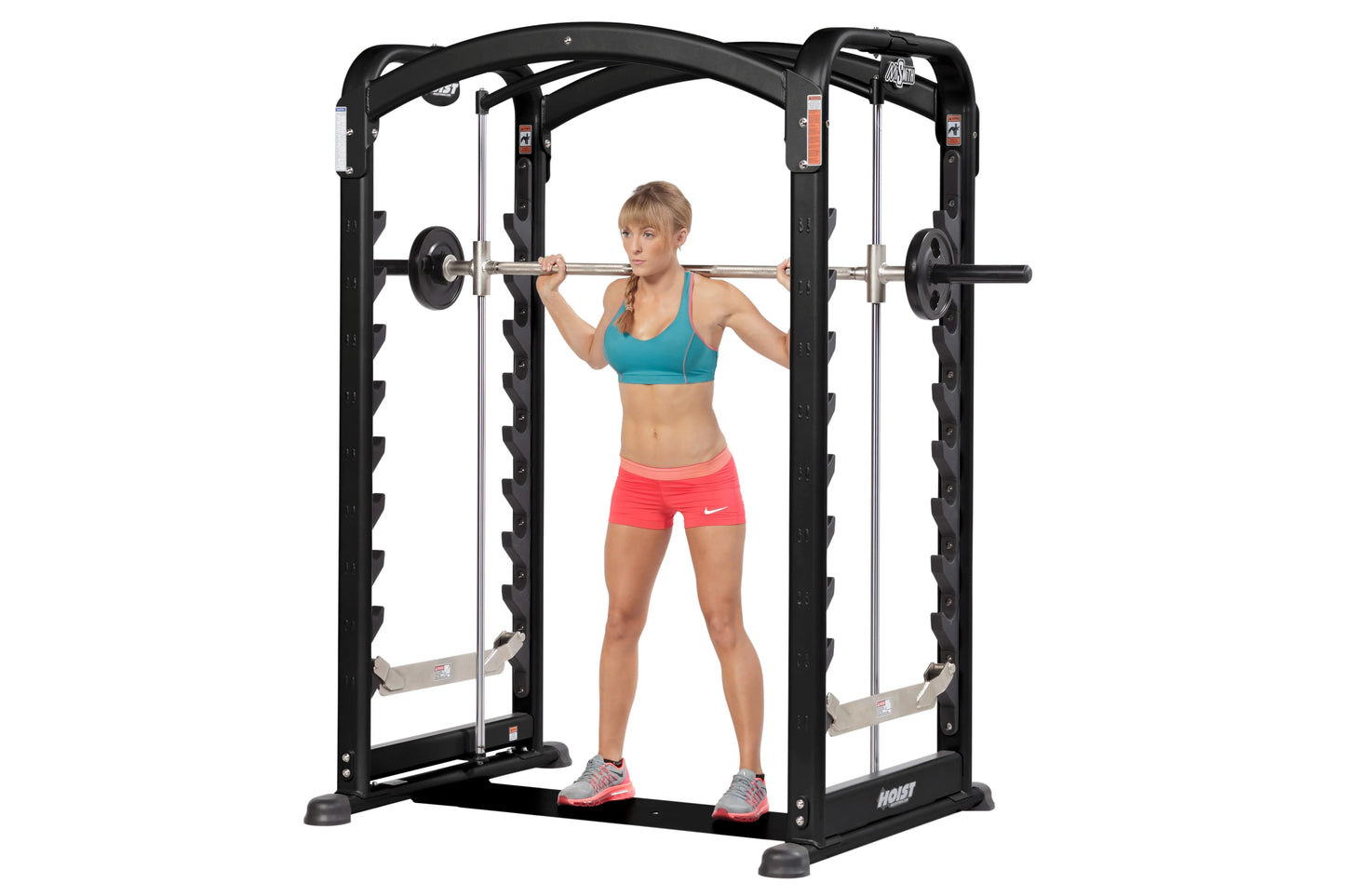 Black Hoist Smith machine with dual action arms, woman in teal sports bra and coral shorts using barbell, white background