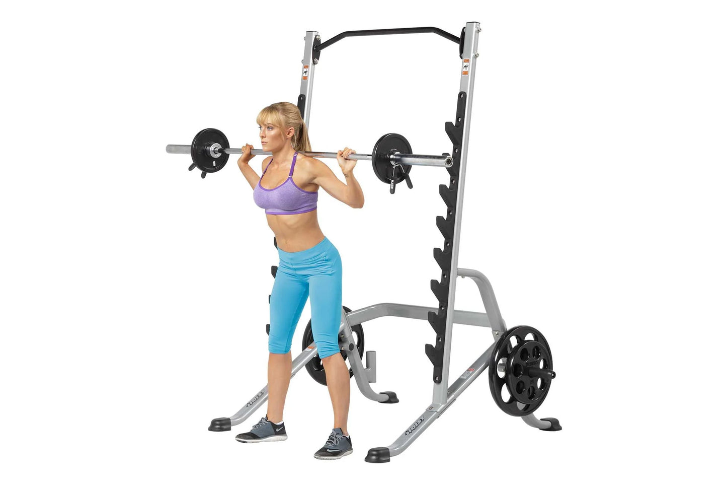Woman using silver and black squat rack with barbell and weight plates, wearing purple sports bra and blue capri leggings