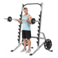 Man wearing light blue tank top and black shorts using silver and black squat rack with barbell and weight plates on white ba