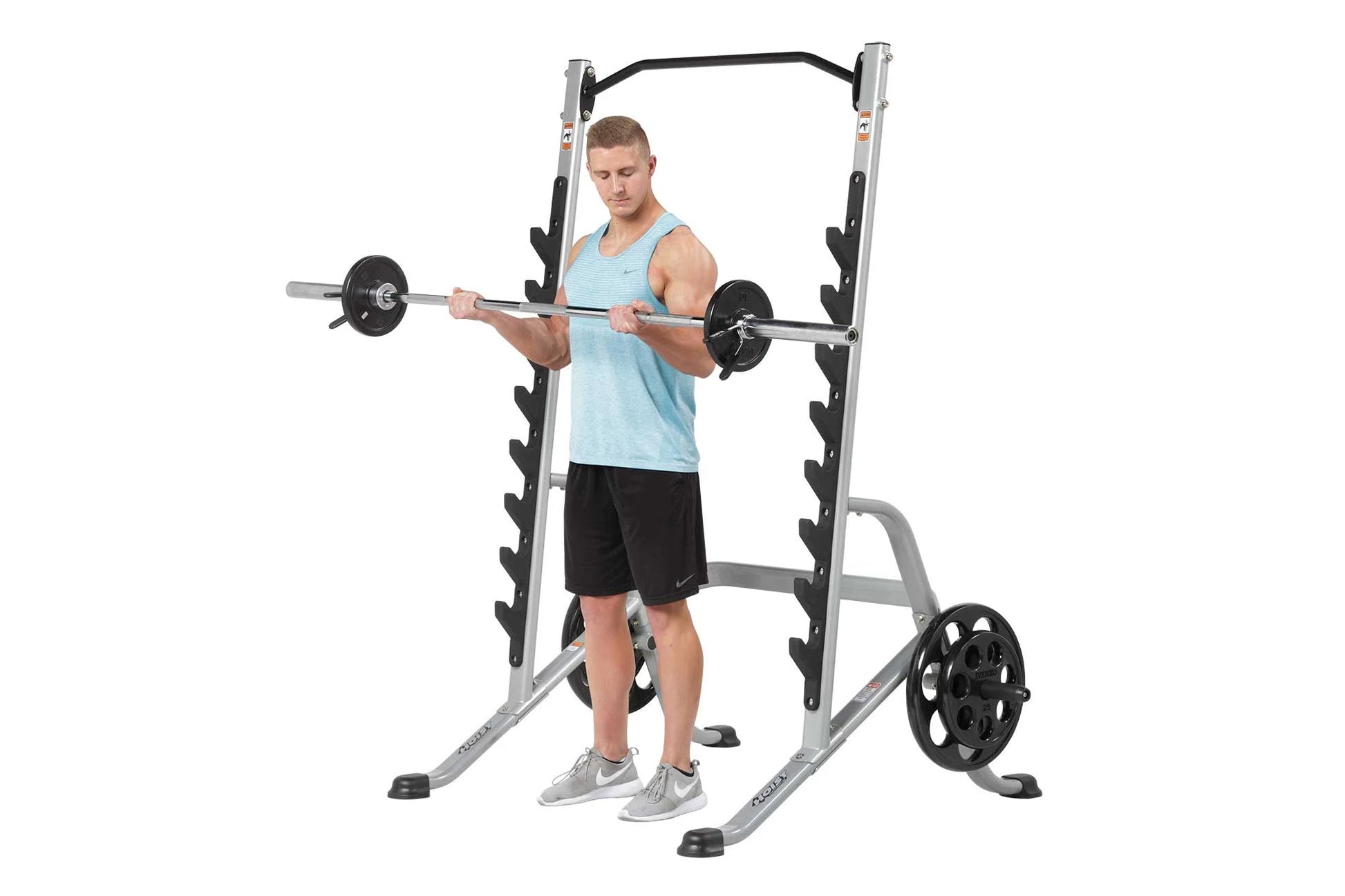 Man wearing light blue tank top and black shorts using silver and black squat rack with barbell and weight plates on white ba