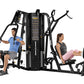Black multi-station gym equipment with two users demonstrating exercises, featuring stacked weight plates and instruction pan