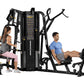 Black multi-station gym equipment with two users demonstrating exercises, featuring weight stacks and instruction panels on w